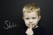 image of infants  - Smart young boy wearing a navy blue jumper stood infront of a blackboard with his finger over his lips being quiet - JPG 