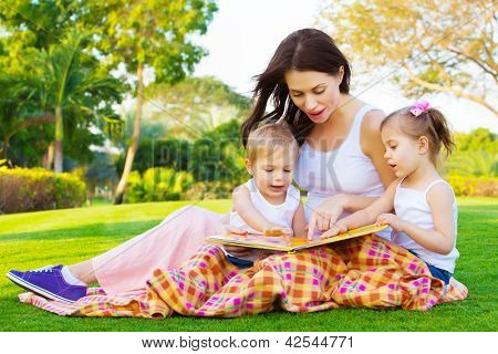 Picture or Photo of Photo of young brunette woman teaching two sweet kids, cheerful mother with cute daughter and lovely son read interesting fairytale, children enjoying book on backyard in daycare, spring season