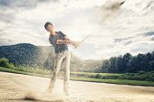 picture of golf  - Male golf player in blue shirt and grey pants hitting golf ball out of a sand trap with sand wedge and sand caught in motion - JPG 