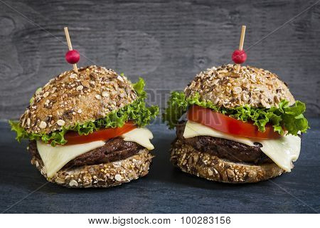 Two gourmet hamburgers with swiss cheese and fresh vegetables on multigrain buns over dark background