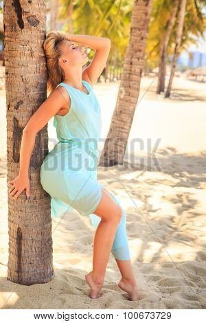 Blonde Girl In Transparent Frock Leans On Palm Trunk