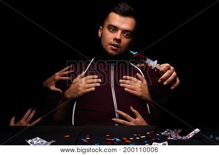 Many hands coming from the dark instigating a depressed student to try a syringe with abusive drugs. A disappointed in life male examines many drugs in capsules and tablets on the black background.