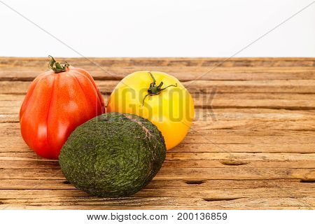 Two heirloom tomatoes and an avocado in a grouping on a cutting board with white background. One tomato is red and one is yellow.