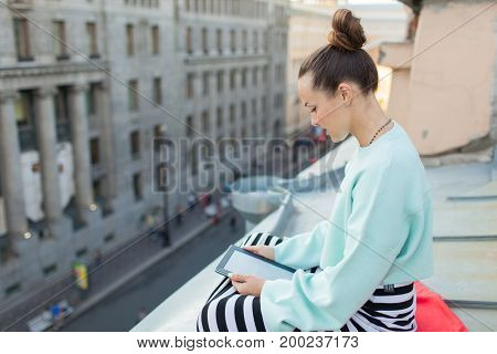 Attractive And Stylish Girl Sits On The Roof Of The House In The Old Town And Reads The E-book