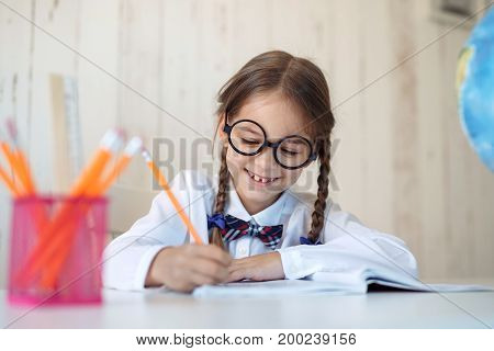 Hard-working little schoolgirl with two pigtails, wearing big glasses and school uniform, doing her homework while sitting at table, writing in copy book with happy expression. Children, education
