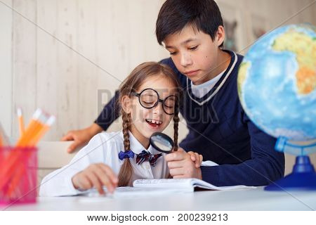 Handsome schoolboy showing his little sister lens. Pretty small girl in big glasses and school uniform, having fun while reading book with big magnifying glass, surrounded with school accessories