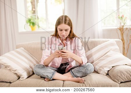 Front view of young girl communocating by cell phone. Female at home, sitting on sofa, writing sms.