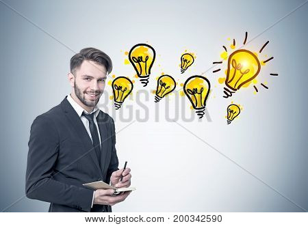 Portrait of a smiling young bearded businessman in a black suit and a tie holding a planner and a pen and looking at the viewer. He is standing near a gray wall with many yellow light bulbs