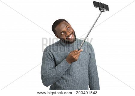 Handsome man taking photo with selfie stick. Beard afro american smiling guy taking picture with phone on monopod. Positive man in grey sweater with monopod on white background.