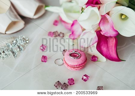 Wedding rings on the pink macaron with pink gems, shoes and flowers background