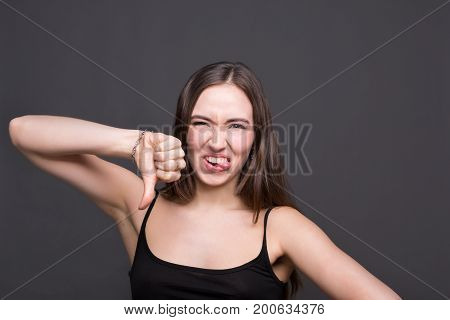 Young woman grimacing at camera, specifies with thumb down. Pretty girl shows tounge, dark background