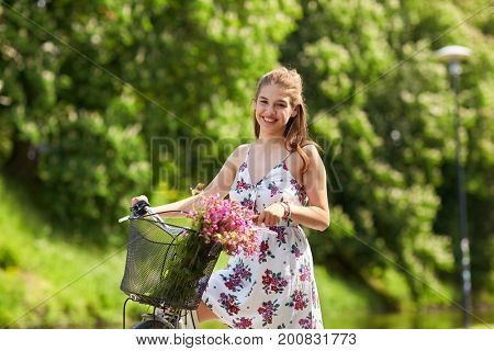 people, leisure and lifestyle - happy young hipster woman wearing summer dress riding fixie bicycle with wild flowers in basket at park