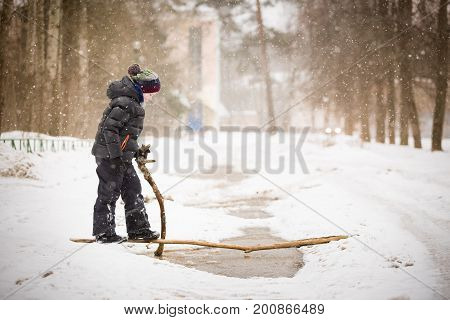 Portrait of cute kid boy with wooden stick trying to cross big puddle on a winter day. Child walking ourdoors and exploring nature. Lifestyle concept