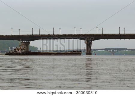 Cargo Delivery By River Transport. The Tug Boat Towing A Barge With Sand. Paton Bridge And Metro Bri