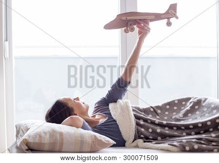 Happy Young Woman Lying In Bed And Holding Wooden Airplane Toy In Air And Daydreaming