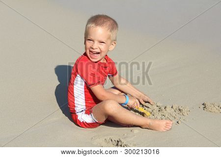 A Little Blond Boy In Clothes With Uv Filter Is Playing With Sand On The Beach By The Sea, Holiday W