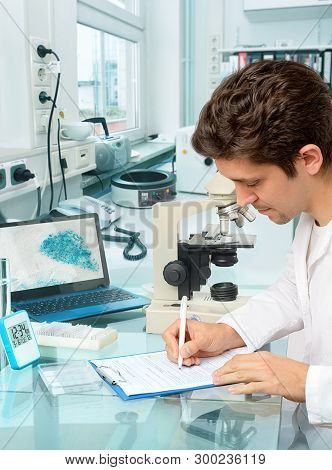 Young Male Scientist Or Tech Makes Notes In Histopathology Laboratory With Light Microscope And A La
