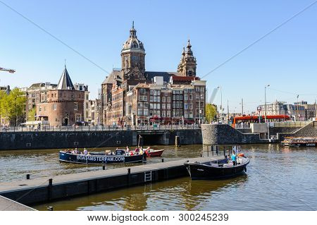 Amsterdam, Netherlands - 19 April 2019: Tourists sightseeng at Canal Boats near the Central Station of Amsterdam. View to Basilica of Saint Nicholas