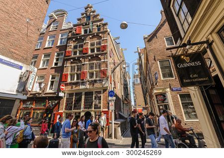 Amsterdam, Netherlands - 19 April 2019: People walking at streets of historic center of the city at spring time, leaning forward facades with red shutters are typical for local houses