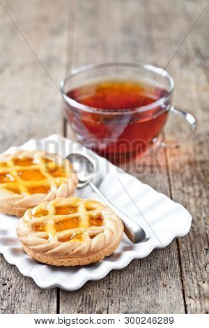 Fresh baked tarts with marmalade or apricot jam filling on white ceramic plate and cup of tea on rustic wooden table background.