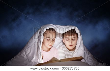 Engrossed Little Girl And Boy Reading Book In Bed After Bedtime. Pretty Kids Together Hiding Under B