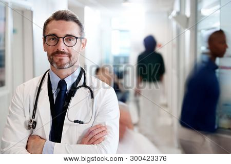 Portrait Of Mature Male Doctor Wearing White Coat With Stethoscope In Busy Hospital Corridor