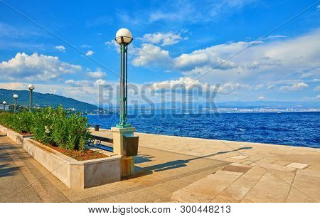 Lovran, Croatia. Sea embankment. Coast of Adriatic Sea. Sunny summer landscape with blue sky and clouds. Green bushes, bench and street lamps along street.