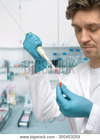 Young Male Scientist Or Graduate Student In Lab Coat Takes An Aliquote Of Liquid Sample. Focus On Th