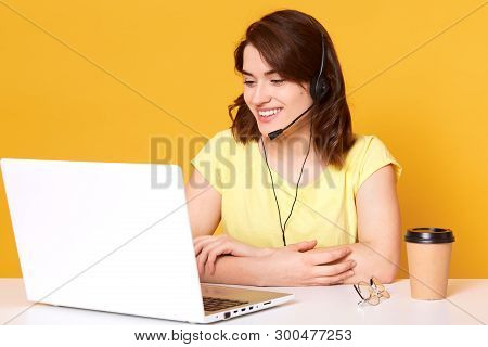 Smiling Energetic Female Sitting At Office Desk With Folded Arms, Having Headset, Looking At Laptop 