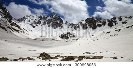 Panoramic View On Plateau In High Snowy Mountains With Frozen Lake Covered Snow At Sunny Spring Day.