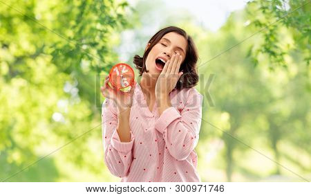 people and bedtime concept - sleepy young woman in pajama with alarm clock showing half past six yawning over background