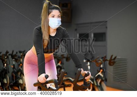 Young Caucasian Girl With A Face Mask On A Stationary Bike In An Empty Gym Due To The Low Capacity I