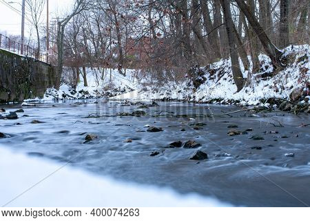 A Long Exposure Of A Small Flowing Creek Next To A Snow Covered Bank With Trees And Foliage