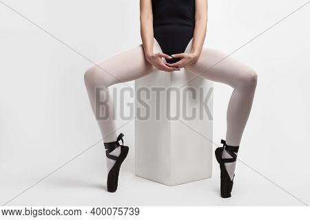 Legs Of Caucasian Ballet Dancer In Body Suit And Pointes Shoes Sitting On Cube Against White Backgro