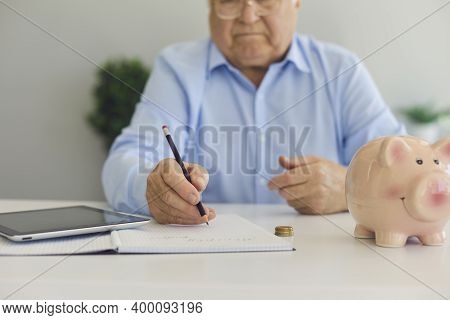 Elderly Man In Shirt In Glasses Sitting With Calculator And Piggybank For Savings And Making Notes