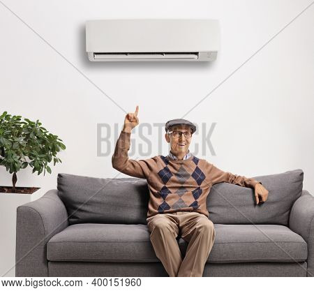 Elderly man seated on a sofa pointing up towards an air conditioning unit on the wall 