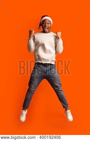 Overjoyed Black Guy In Santa Hat Celebrating Success, Jumping Over Orange Background With Raised Fis