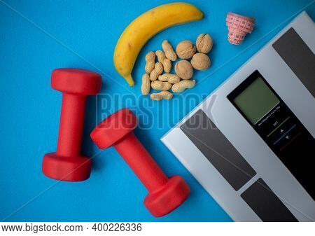 Electronic Scale, Banana, Nuts, Measuring Tape And Dumbbells On A Blue Background. Food And Fitness 