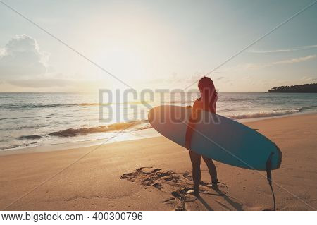 Woman Hold Surfboard Standing At Tropical Sunset Beach Background. Summer Vacation And Sport Adventr