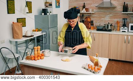 Woman Baker Sieving Flour On Bread Dough With Metallic Sieve On Table In Kitchen. Happy Elderly Chef