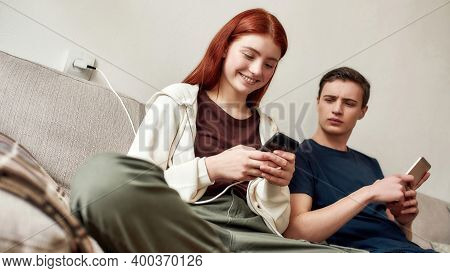 Couple Of Teenagers Sitting On The Couch At Home, Using Mobile Phone. A Guy Jealously Looks At Girlf