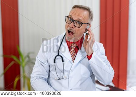 Funny Doctor With Good Sence Of Humor Poses In Medical Room. Modern Senior Medic In Scrubs.
