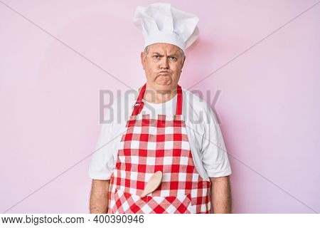 Senior grey-haired man wearing professional baker apron puffing cheeks with funny face. mouth inflated with air, crazy expression. 