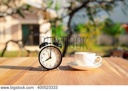 Black Alarm Clock And White Cup Coffee On Wooden Table In The Morning