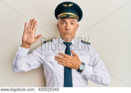 Handsome middle age mature man wearing airplane pilot uniform swearing with hand on chest and open palm, making a loyalty promise oath 