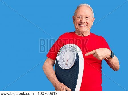 Senior handsome grey-haired man holding weighing machine smiling happy pointing with hand and finger 