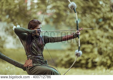 Woman With Arrows And Bow Sits On A Fallen Tree And Takes Aim At A Target