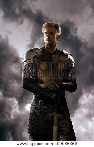 Ancient knight in metal armor with sword standing on a cloudy background