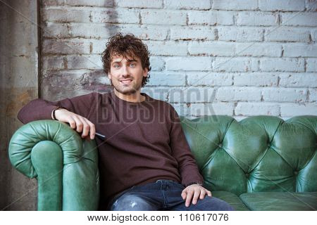 Attractive smiling joyful pleased content positive curly confident male in brown sweetshirt and jeans sitting on green leather couch
