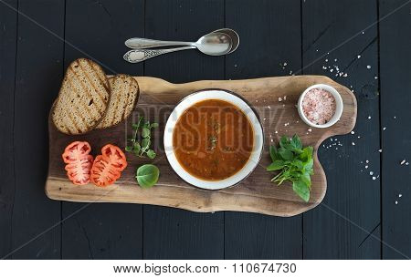 Roasted tomato soup with fresh basil, spices and bread in vintage metal bowl on wooden board over bl